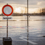 Ein Straßenschild mit dem Zusatz "Hochwasser" steht vor einer Wasserfläche im Abendlicht.