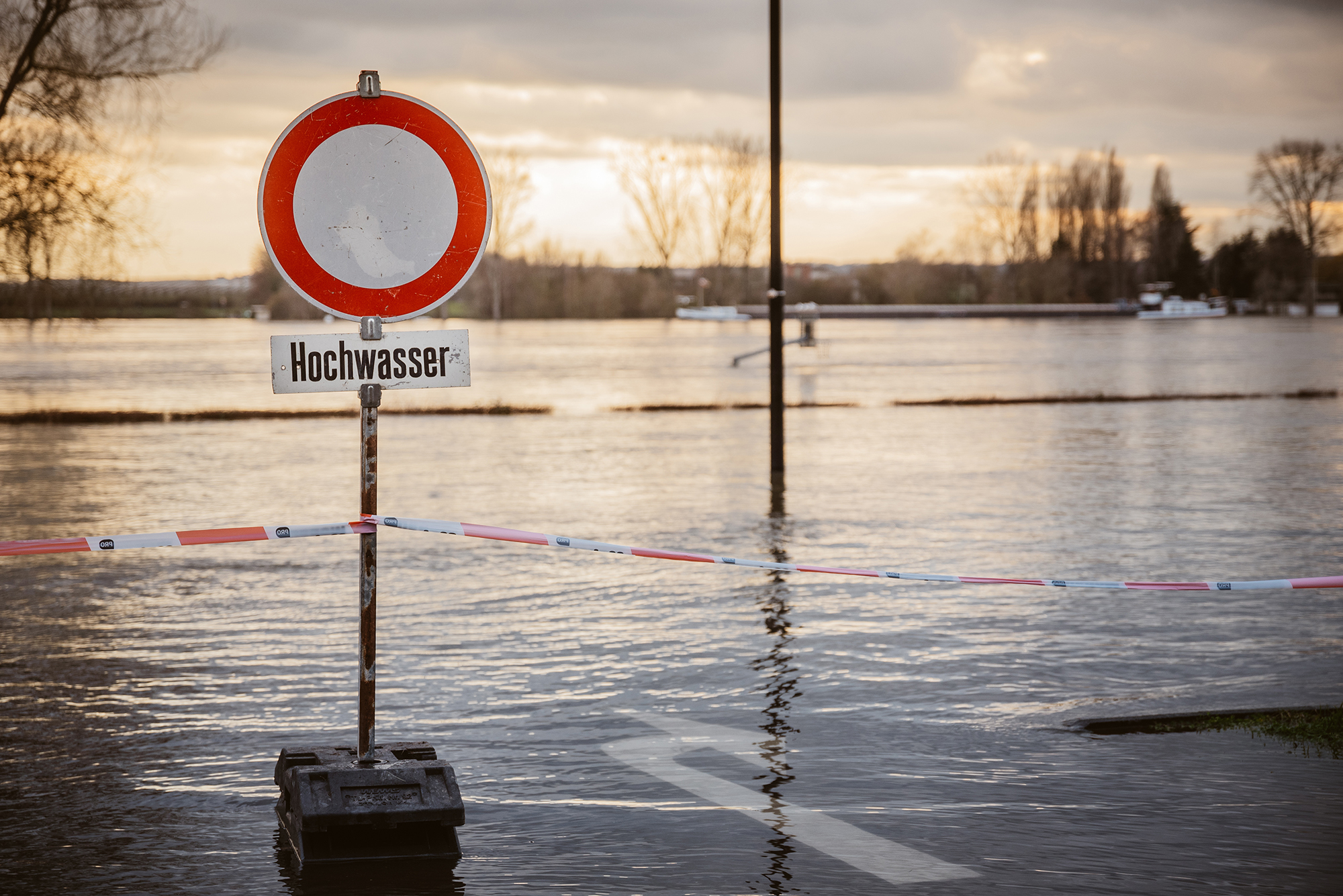 Ein Straßenschild mit dem Zusatz "Hochwasser" steht vor einer Wasserfläche im Abendlicht.