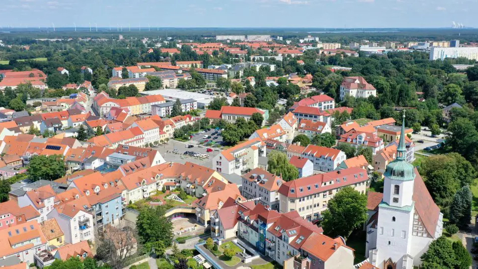 Luftaufnahme der Altstadt von Hoyerswerda, vorwiegend historische Häuser mit roten Ziegeldächern, im Hintergrund Bäume, im Vordergrund ein heller Kirchturm.