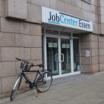 Straßenszene, graurötliche Hausfassade mit großer gläserner Eingangstür, davor ein geparktes Fahrrad. Über der Tür ein großes Schild mit der Aufschrift "Joccenter Essen"