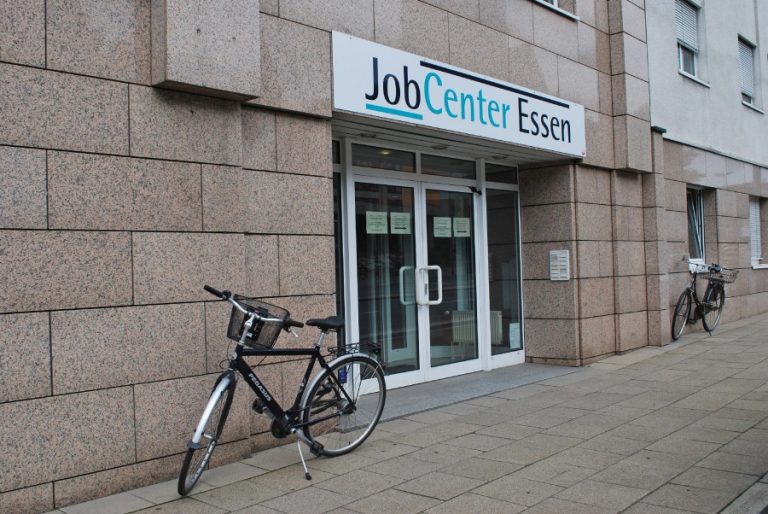 Straßenszene, graurötliche Hausfassade mit großer gläserner Eingangstür, davor ein geparktes Fahrrad. Über der Tür ein großes Schild mit der Aufschrift "Joccenter Essen"