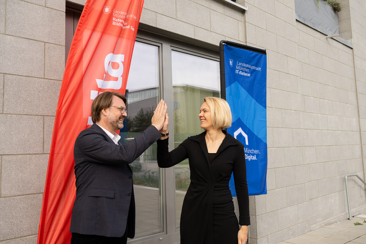 Ein Mann und eine Frau in dunkler, förmlicher Kleidung stehen vor der gläsernen Tür eines modernen Gebäudes und geben sich "High Five"