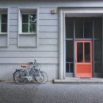 Streng geometrische Hausfassade mit klassizistischen Elementen, ganz in grau, nur die Haustür ist Orange. Davor parken zwei Fahrräder.