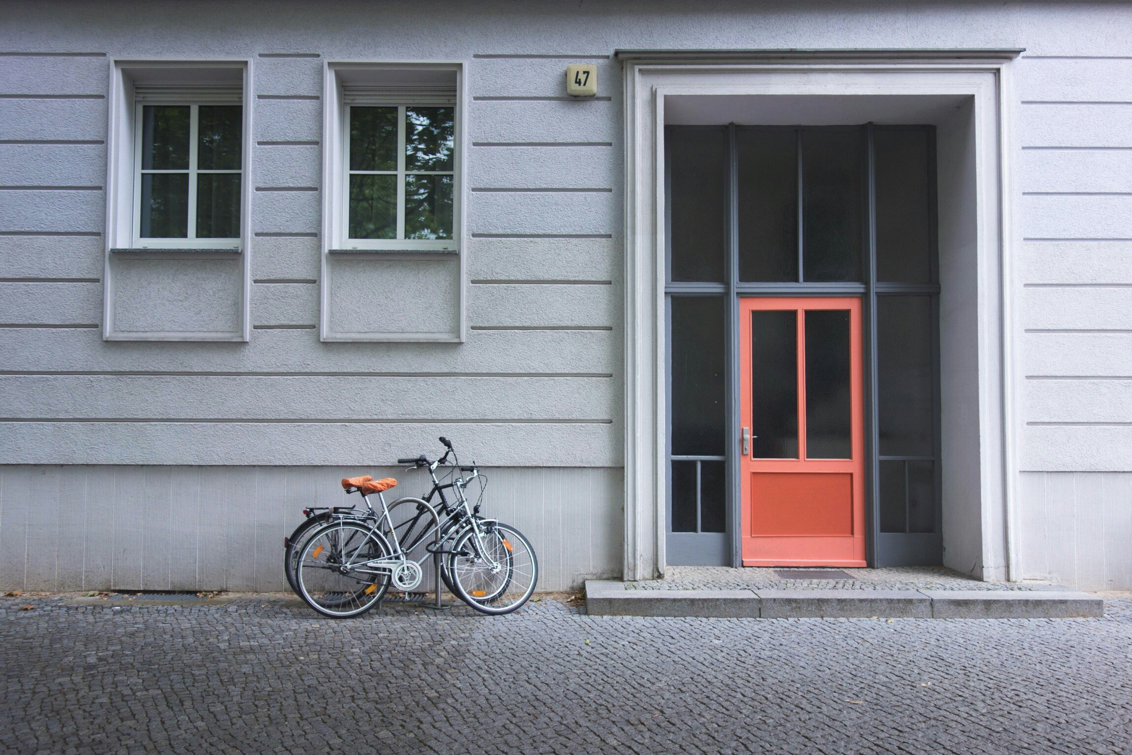 Streng geometrische Hausfassade mit klassizistischen Elementen, ganz in grau, nur die Haustür ist Orange. Davor parken zwei Fahrräder.