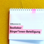 Auf einem gelben Polsterstoff (vermutlich von einem sessel) liegt ein gedruckter Zettel mit der Aufschrift: "Willkommen zum Reallabor Bürger*innen-Beteiligung"