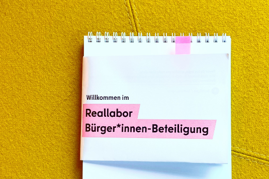 Auf einem gelben Polsterstoff (vermutlich von einem sessel) liegt ein gedruckter Zettel mit der Aufschrift: "Willkommen zum Reallabor Bürger*innen-Beteiligung"