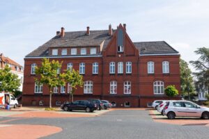 Zweistöckiges Gebäude aus rotem Backstein mit Dachgauben, davor ein Parkplatz mit wenigen Autos und spärlicher Bepflanzung.