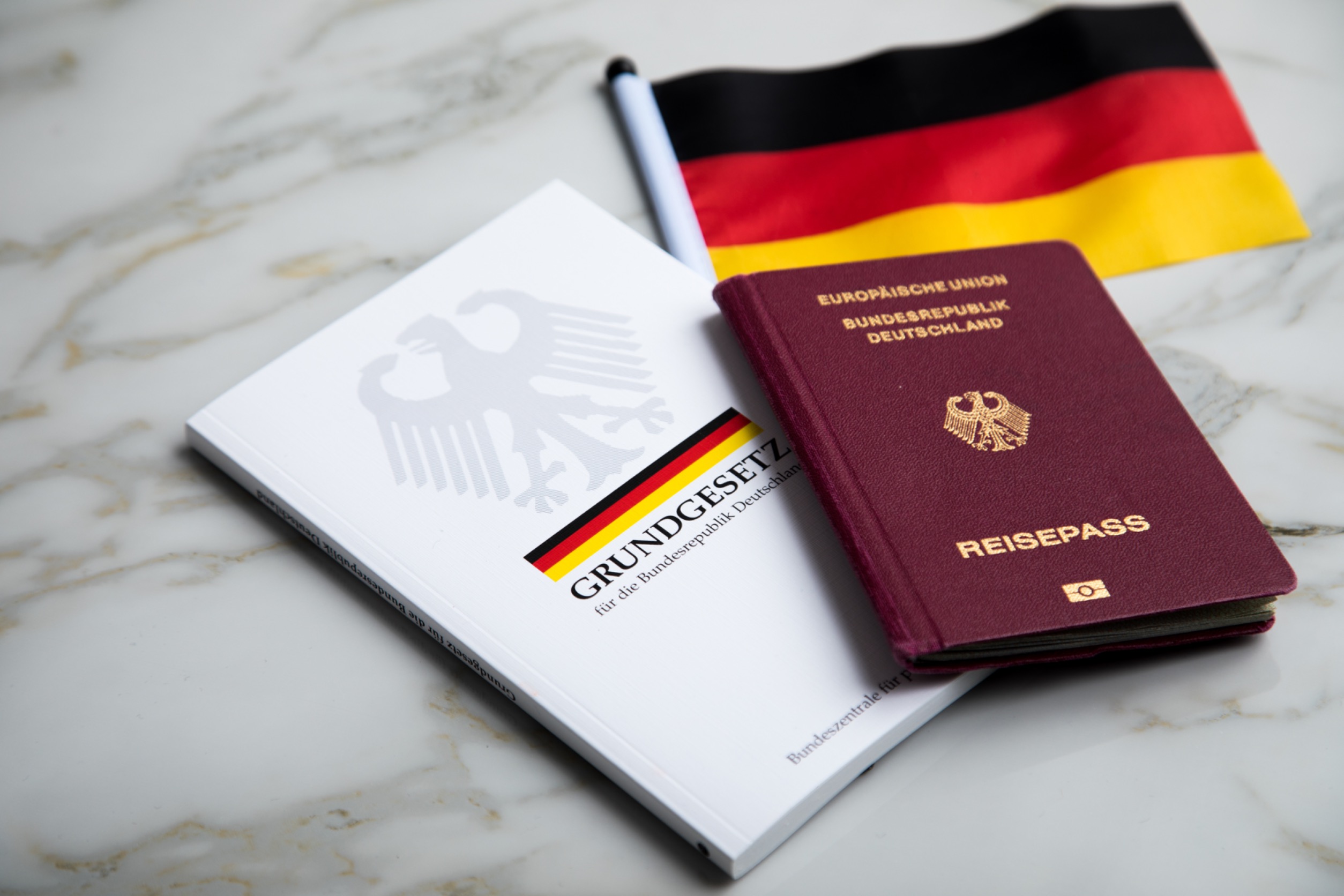 Ein deutscher Reisepass, das deutsche Grundgesetzbuch und eine Deutschlandflagge liegen auf einer marmorierten Unterlage.