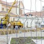 Baustelle: Blick durch Zaundraht auf einen Pumpenkran zum Heben und Gießen von Beton, im Hintergrund ein Gebäude, das eine Schule sein könnte.