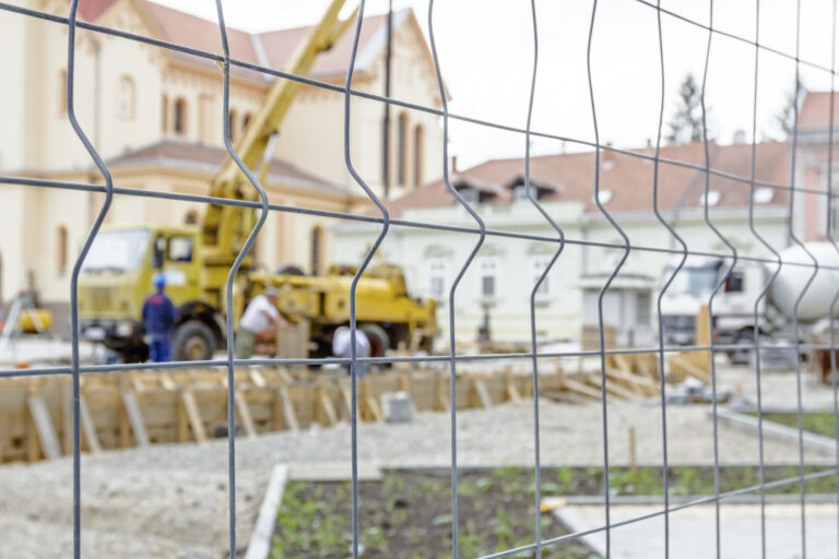 Baustelle: Blick durch Zaundraht auf einen Pumpenkran zum Heben und Gießen von Beton, im Hintergrund ein Gebäude, das eine Schule sein könnte.