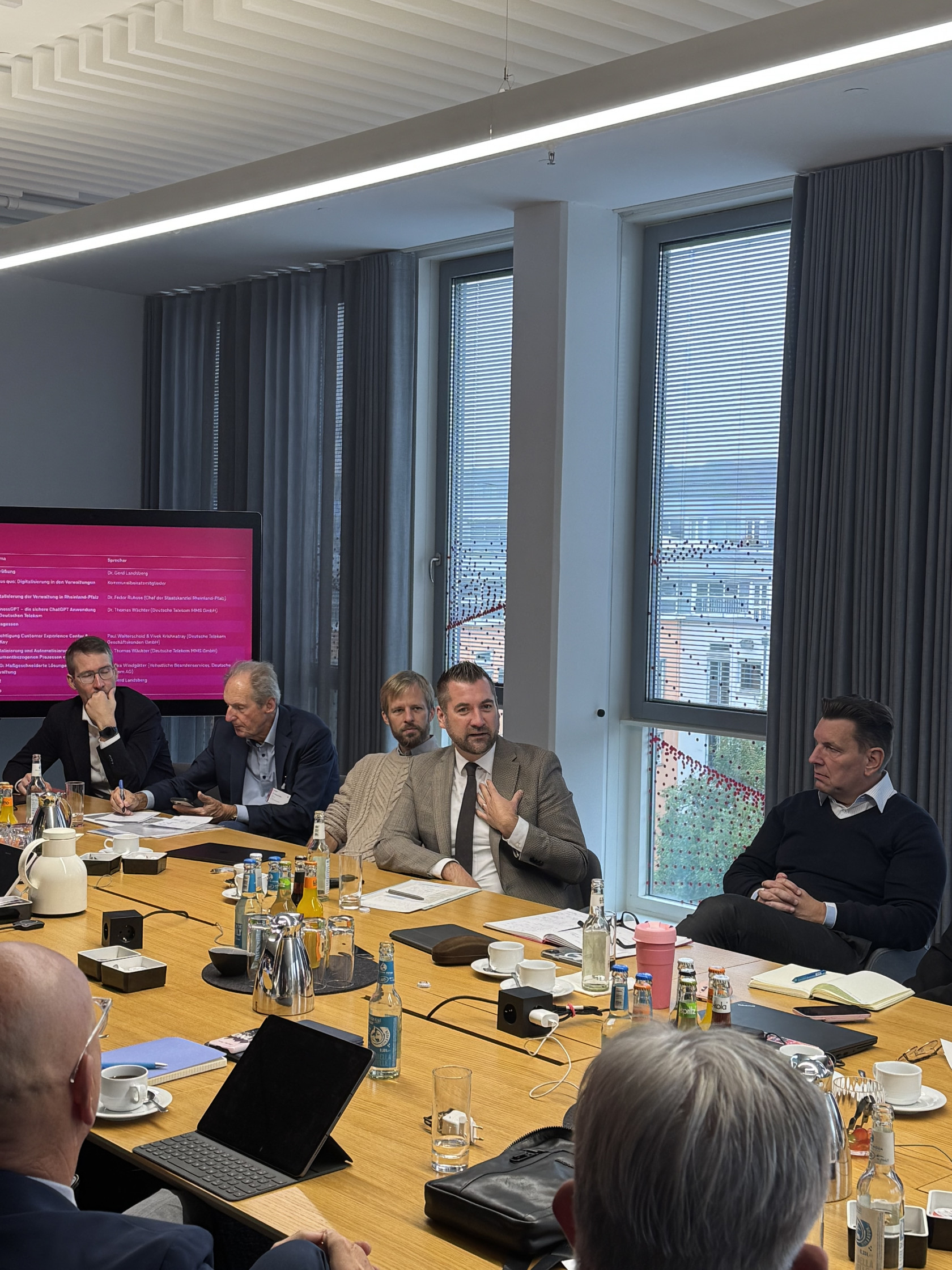 Ein Sitzungssaal mit Konferenztisch aus hellem Holz, daran sitzen Menschen mit förmlicher Kleidung, im Hintergrund ein Bildschirm mit magentafarbener Anzeige.