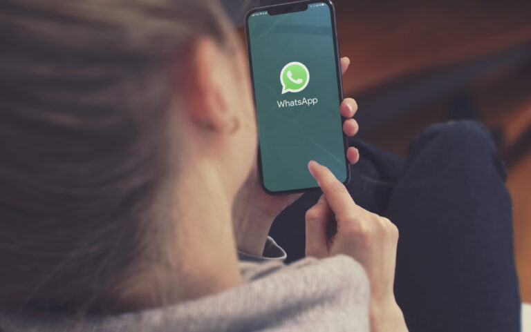 Man sieht von hinten über die Schulter einer jungen Frau, die auf ihr Smartphote tippt, dort ist das WhatsApp-Logo zu sehen.