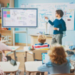 Schulkinder sitzen im Klassenzimmer an ihren Tischen und blicken auf ein Whiteboard, an dem ein Lehrer etwas erklärt.