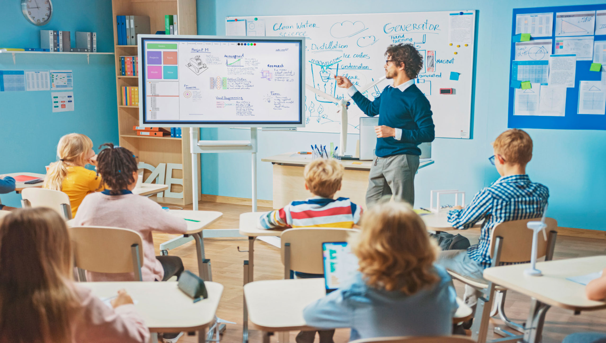 Schulkinder sitzen im Klassenzimmer an ihren Tischen und blicken auf ein Whiteboard, an dem ein Lehrer etwas erklärt.