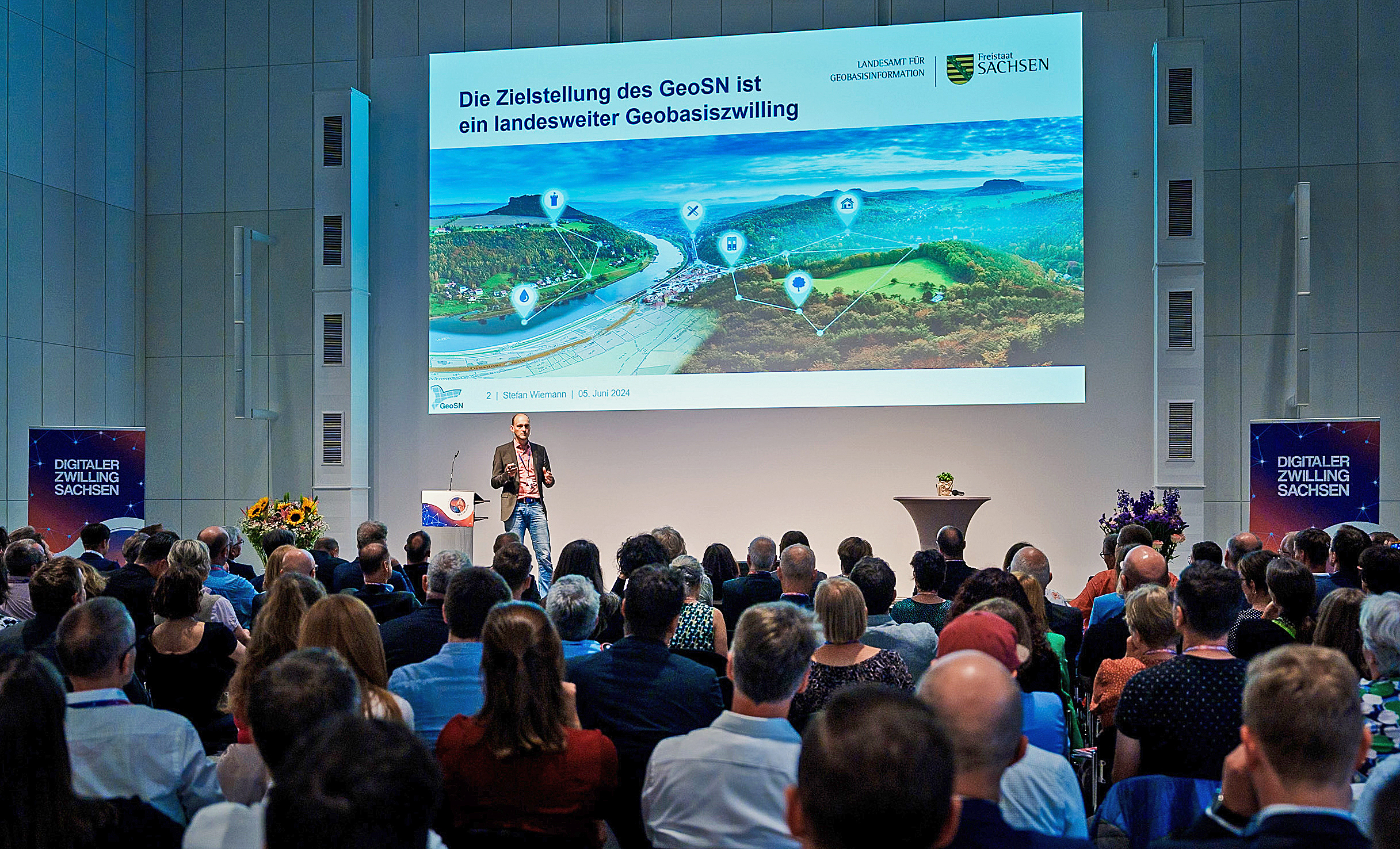 Momentaufnahme der Kick-Off-Veranstaltung, die auf der Bühne einen Referenten zeigt, hinter ihm an die Wand projiziert eine Vortragsfolie, vor der Bühne sitzen zahlreiche Zuhörer, die von hinten zu sehen sind.