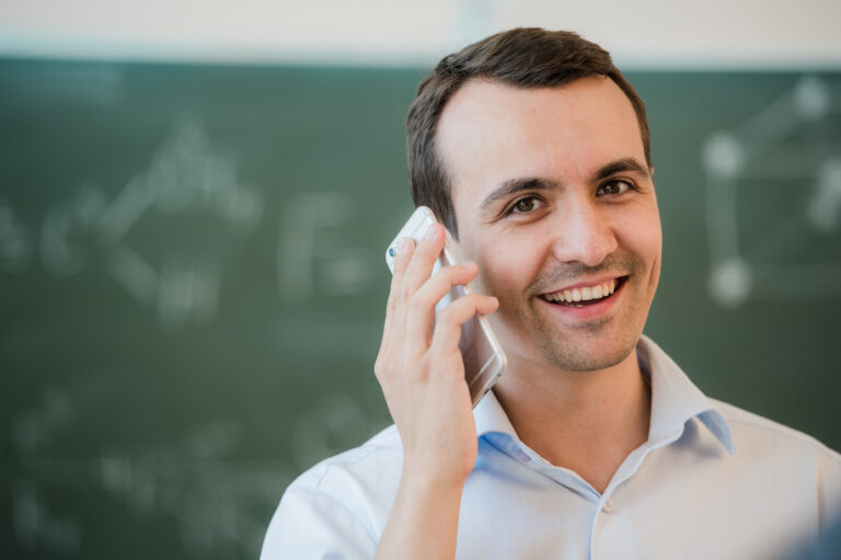 Porträt eines am Smartphone telefonierenden Mannes, im Hintergrund eine grün, mit Kreide beschriebene Tafel.