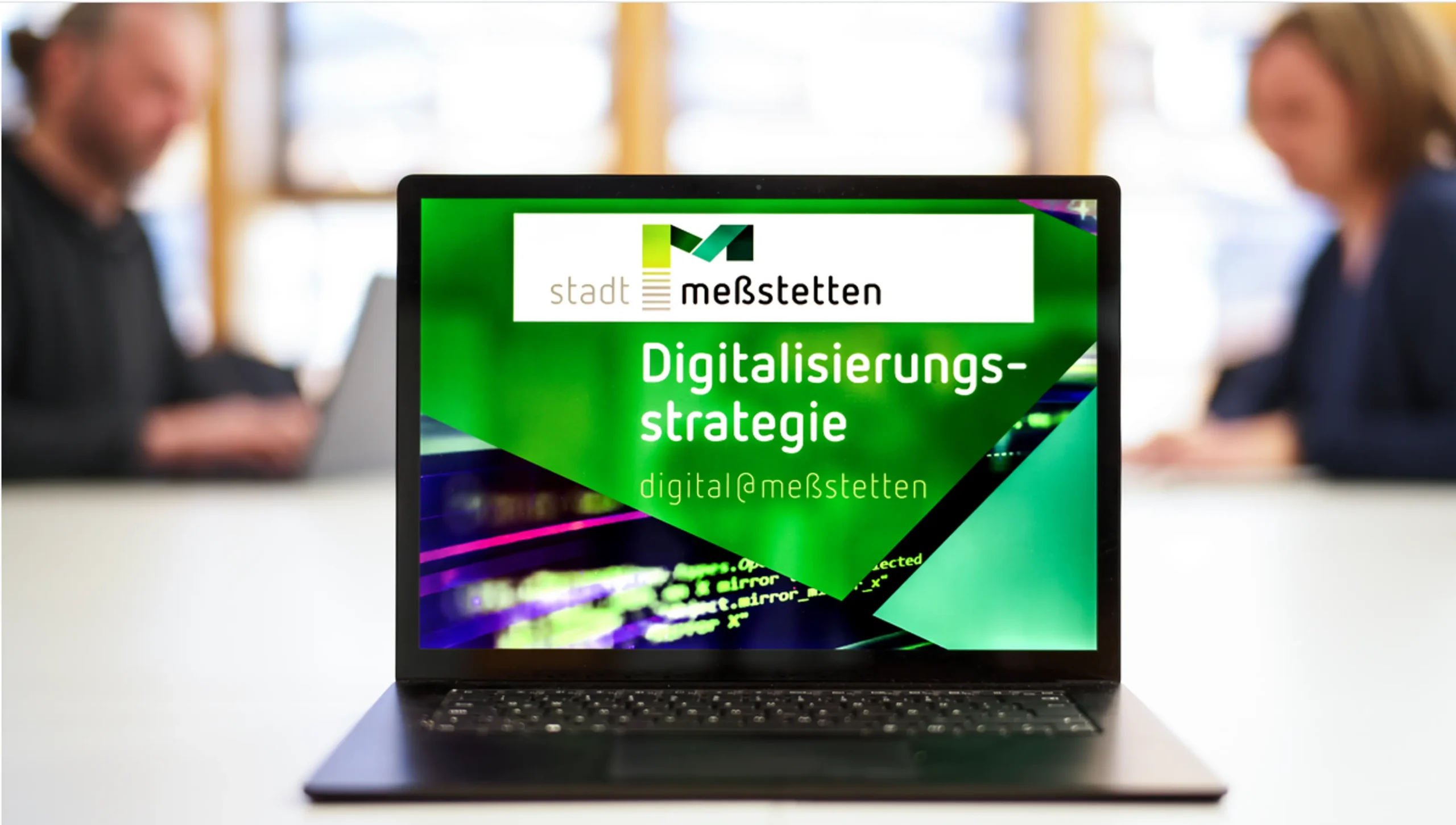 Ein aufgeklappter Laptop steht auf einem Tisch und zeigt auf dem Display das Deckblatt der Digitalisierungsstrategie der Stadt Meßstetten. Im Hintergrund sitzen sich ein Mann und eine Frau am Tisch gegenüber und arbeiten selbst an Laptops.