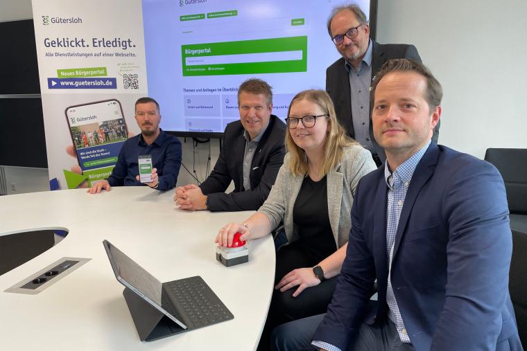 Stephan Junghanns (Online-Redakteur), Carsten Schlepphorst (Beigeordneter für den Bereich Digitalisierung und Organisation), Anna-Lena Hoffmann (Projektleiterin im Fachbereich Digitalisierung), Bürgermeister Matthias Trepper und Thorsten Schmidthuis (Leiter Fachbereich Digitalisierung) sitzen an einem Tisch vor einem aufgeklappten Laptop, im Hintergrund zeigt ein Whiteboard die Startseite des Gütersloher Bürgerportals an.