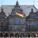 Rathaus der Freien und Hansestadt Bremen, mit Arkaden und drei markanten Stufengiebeln, Deutschlandflagge wehtt, blauer Himmel.