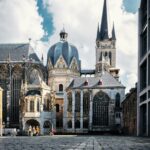 Blick aus leichter Untersicht über eine Kopfsteinpflasterfläche auf den gotischen Aachener Dom, rechts im Vordergrund die Fassade eines modernen Betonbaus.