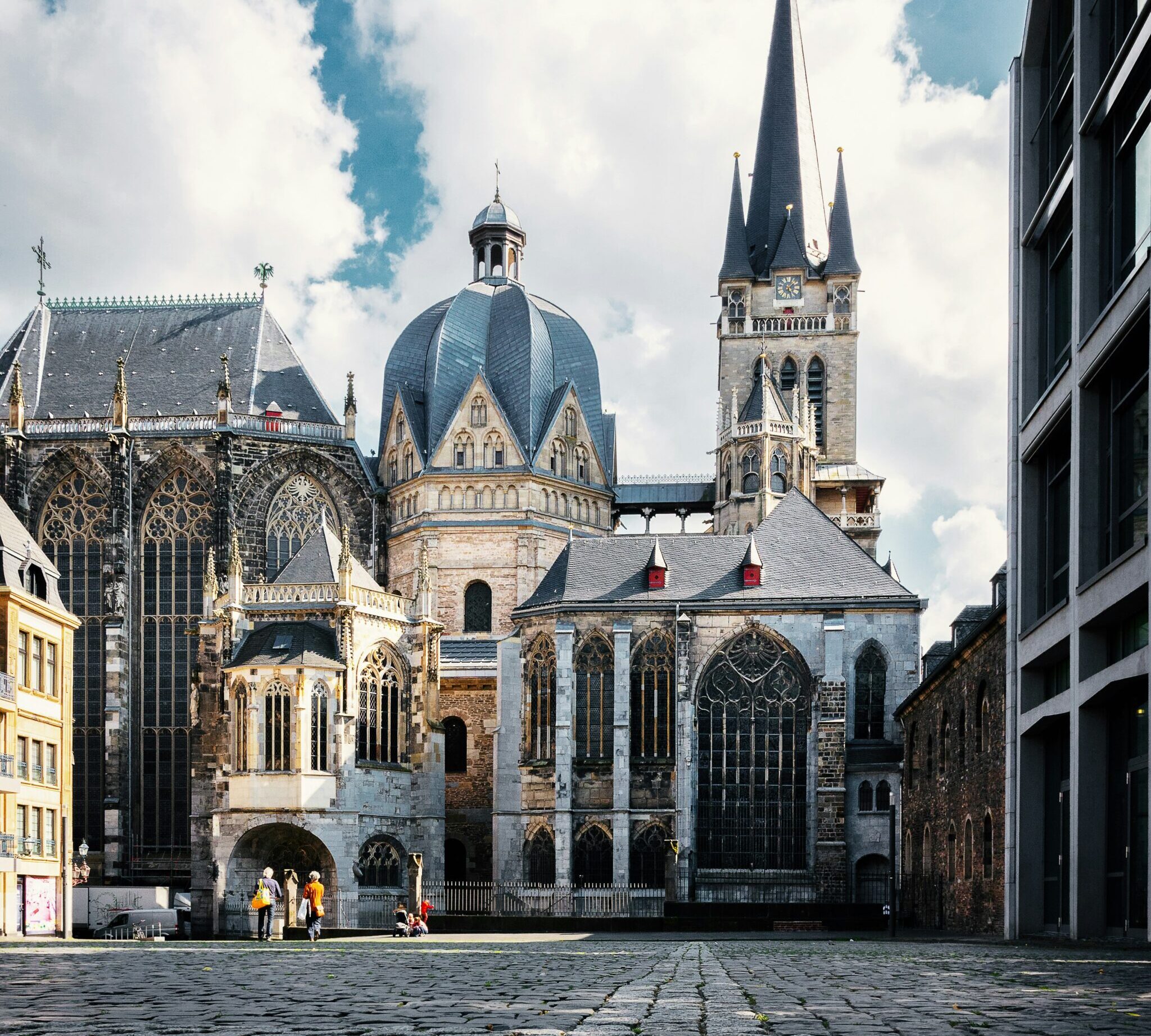 Blick aus leichter Untersicht über eine Kopfsteinpflasterfläche auf den gotischen Aachener Dom, rechts im Vordergrund die Fassade eines modernen Betonbaus.