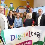 Gruppe von neun personen steht in einem gelb gestrichenen Raum, vor sich ein Transparent mit der mehrfarbigen Aufschrift "Digitaltag".