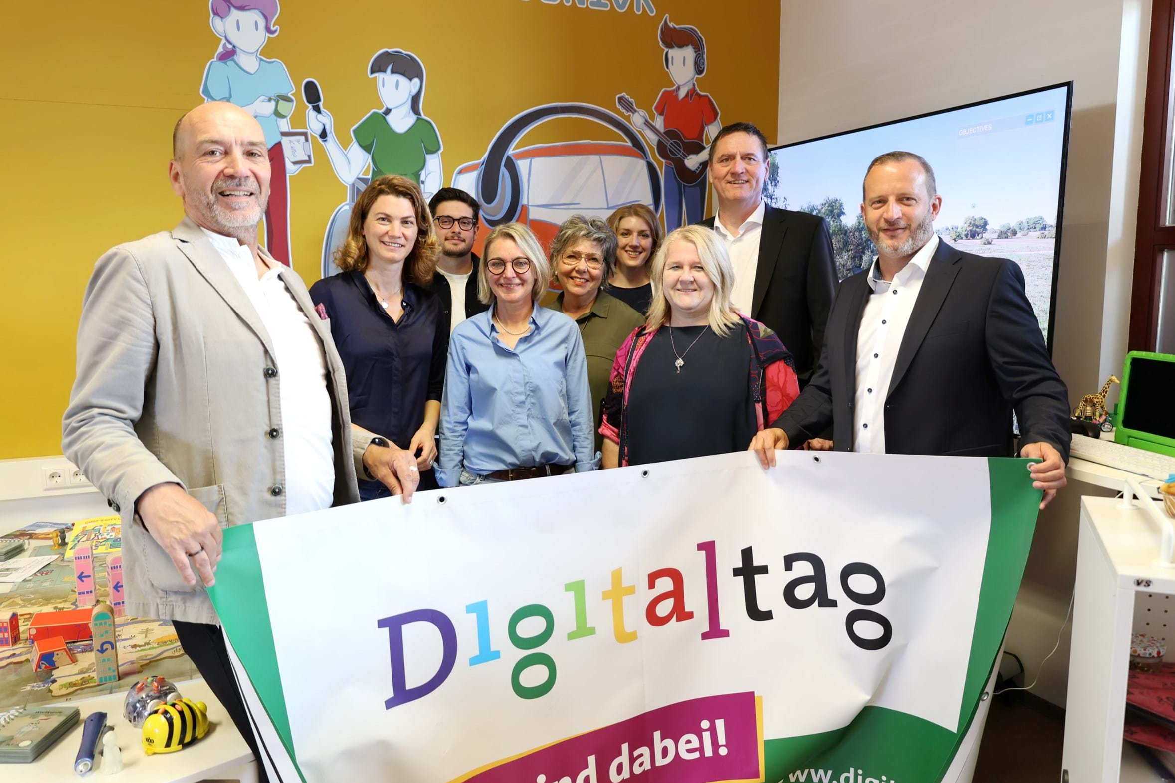 Gruppe von neun personen steht in einem gelb gestrichenen Raum, vor sich ein Transparent mit der mehrfarbigen Aufschrift "Digitaltag".