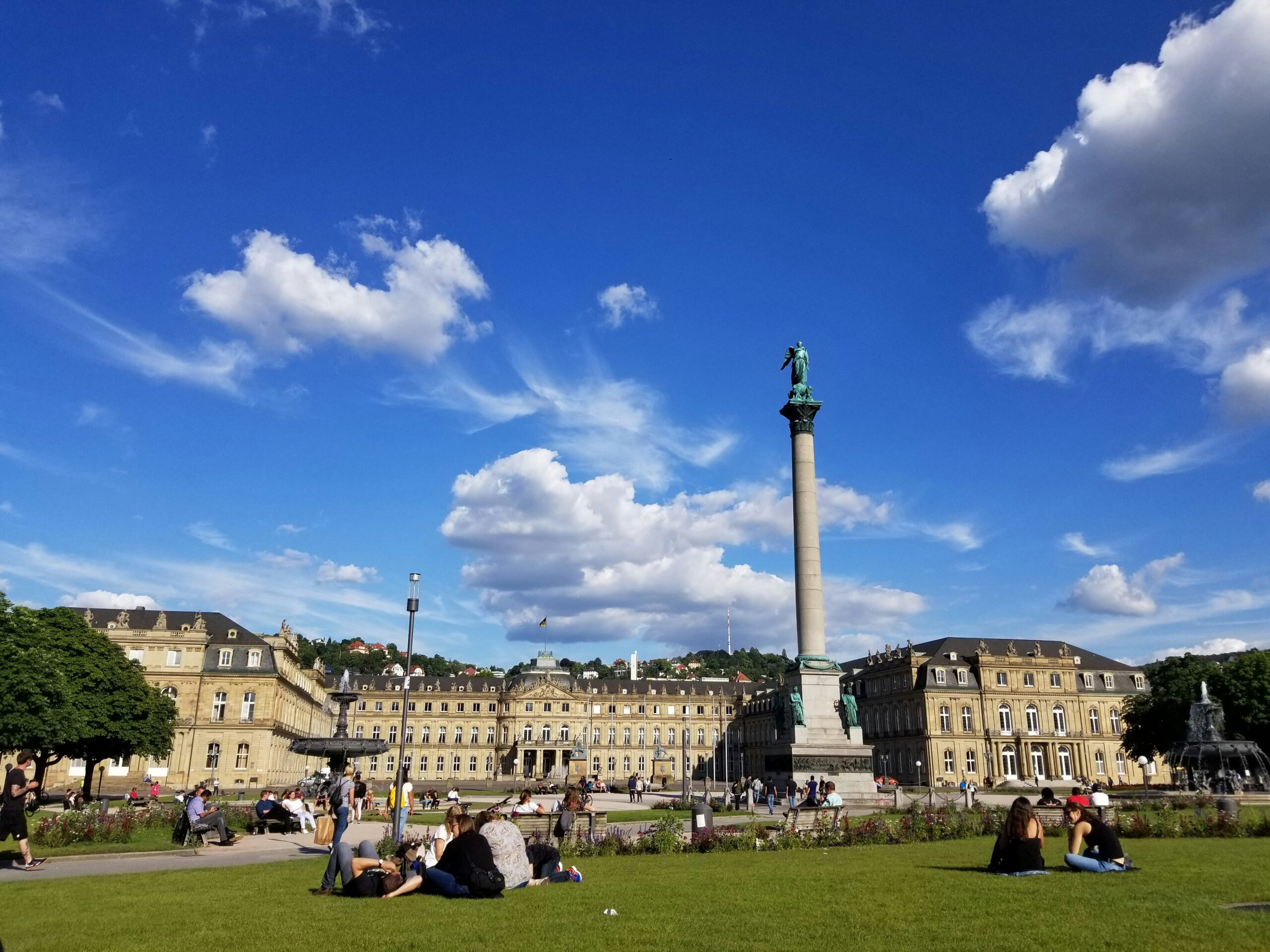 Stuttgarter Schlossplatz mit Jubiläumssäule bei gutem Wetter, Menschen sitzen auf der Wiese.