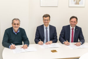 v.l. Roman Starý von Esri, Uwe Hofmann, Schulleiter des Gymnasiums Dresden-Plauen) und GeoSN-Präsident Ronny Zienert sitzen nebeneinander an einem Tisch, jeweils mit Stift in der Hand, um vor ihnen liegende Unterlagen zu unterzeichnen.