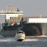 Großes Frachtschiff, davor fährt ein kleines Motorboot.