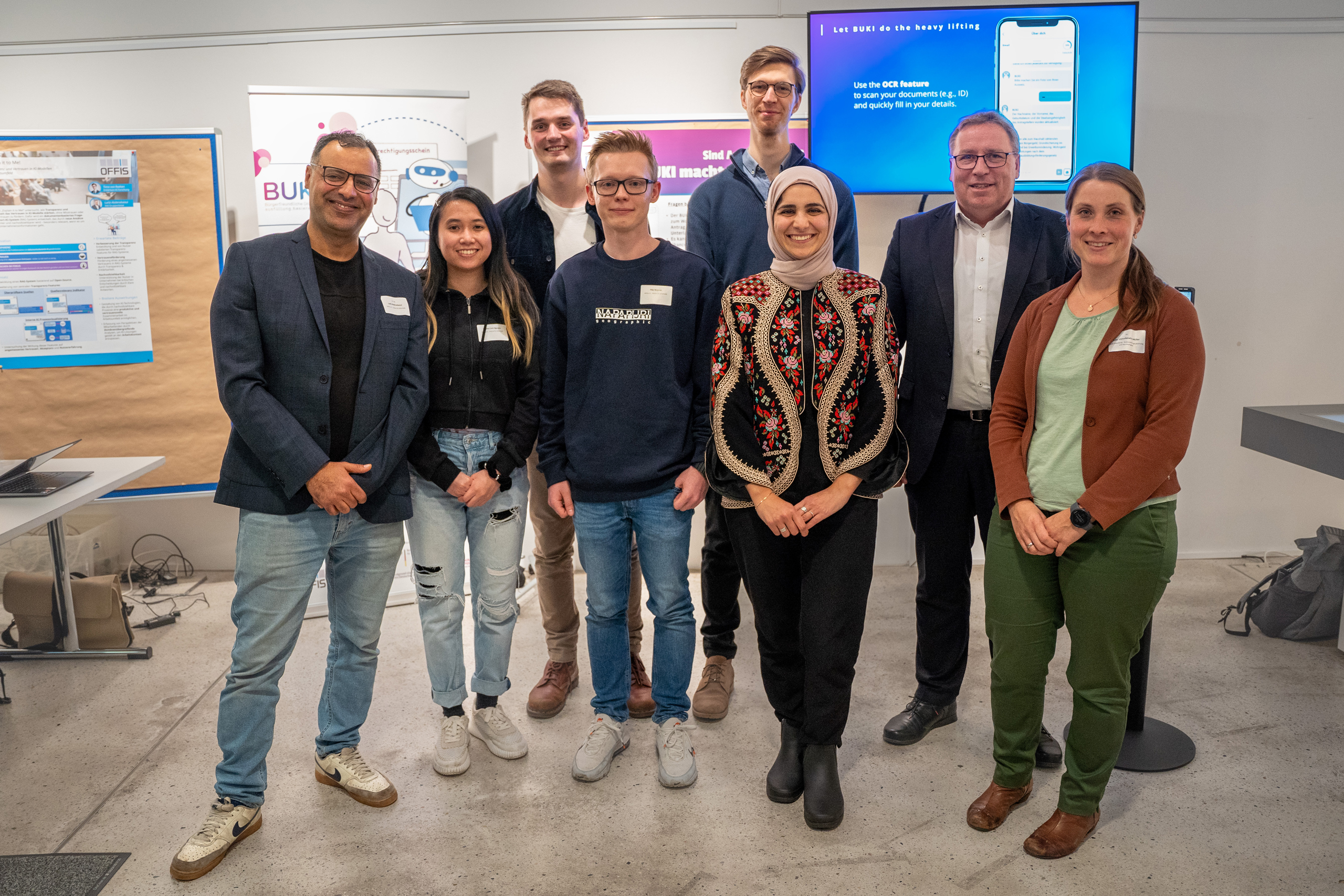 Eine Personengruppe steht in einem Besprechungsraum, im Hintergrund sind unterschiedliche Präsentationsfolien und -Poster rund um das BUKI-Projekt zu sehen.