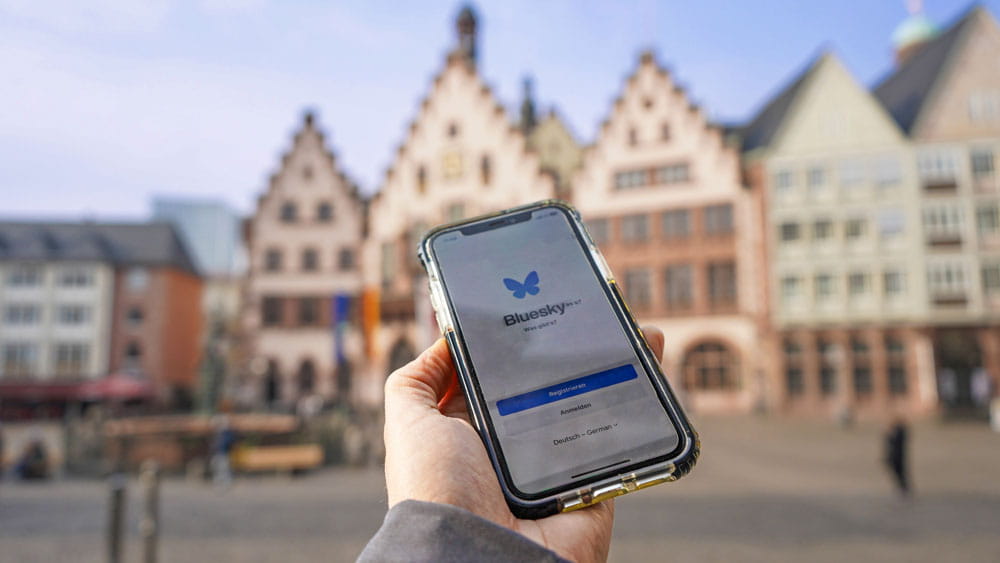 Ein Smartphone zeigt die Bluesky-Oberfläche an, im Hintergrund ist der so genannte Römer in Frankfurt am Main zu sehen.