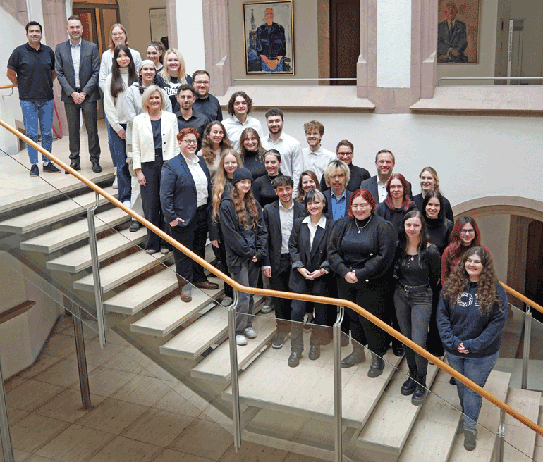 Eine größere Personengruppe mit vornehmlich dunkel gekleideten, jungen Menschen steht in Fotopose auf einer modernen Treppe.