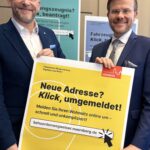 Zwei Männer in Anzug und weißem Hemd präsentieren ein gelbes Plakat, das für die Nutzung der digitalen Wohnungsummeldung dient.