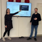 Julia Kleinert vom Team Organisation der Stadt Bad Oeynhausen und Patrick Höwener vom Team Steuern stehen vor einem an der Wand befestigten Whiteboard und erklären darauf den neuen Prozess mit EMMA.