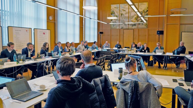 Arbeitsrunde aus etwa 20 Personen mit Laptops an Konferenztischen in einem holzgetäfelten Saal.