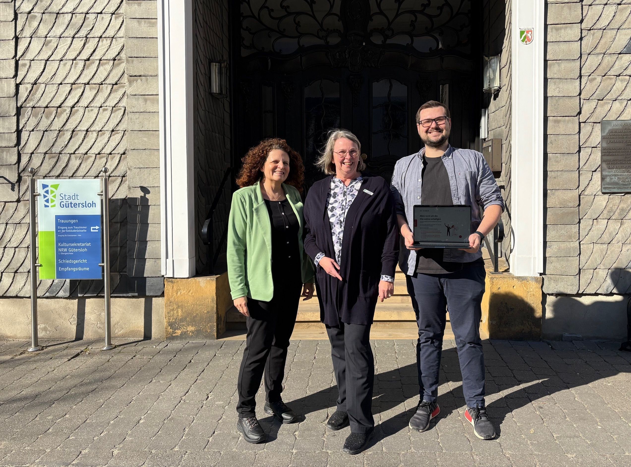 Manuela Marxen, Claudia Wiegelmann und Pascal Stewin stehen nebeneinander vor einem Gebäude, Stewin hält einen aufgeklappten Laptop in der Hand.